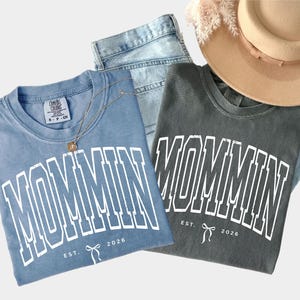 May include: Two t-shirts, one blue and one gray, with the word "MOMMIN" in white block letters. The text "EST. 2026" and a bow graphic are below the word. A pair of blue jeans, a gold necklace, and a tan hat are also in the image.