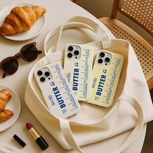 May include: Three phone cases with a butter-themed design are displayed in a cream-colored tote bag. The cases are white and yellow, with the words "SALTED BUTTER AA" and measurement markings printed on them. A croissant, sunglasses, and lipstick are also visible.