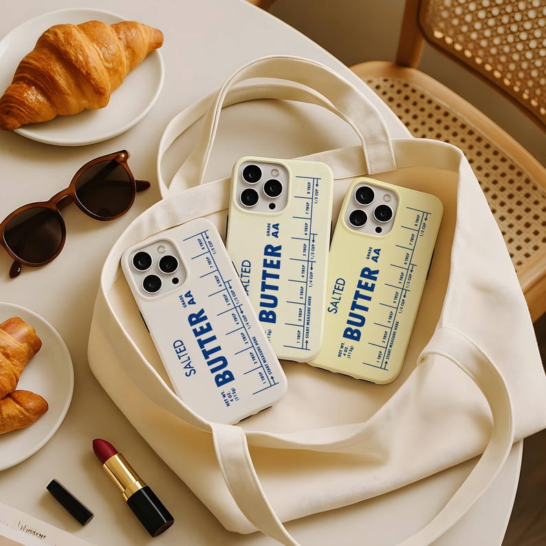 May include: Three phone cases with a butter-themed design are displayed in a cream-colored tote bag. The cases are white and yellow, with the words "SALTED BUTTER AA" and measurement markings printed on them. A croissant, sunglasses, and lipstick are also visible.