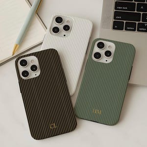 Może przedstawiać: Trzy etui na telefon z pionowymi wzorami w paski. Jedno jest białe, jedno oliwkowozielone z inicjałami "HM", a drugie brązowe z inicjałami "CL". Etui leżą na białej powierzchni z długopisem i klawiaturą laptopa w tle.