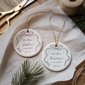 K&ouml;nnte beinhalten: Zwei wei&szlig;e runde Keramik-Weihnachtsornamente mit goldenen Kordelaufh&auml;ngern. Eines hat ein rosa Design mit dem Text "The Year Sophia was born 2025". Das andere hat ein blaues Design mit dem Text "The Year Emerson was born 2025".