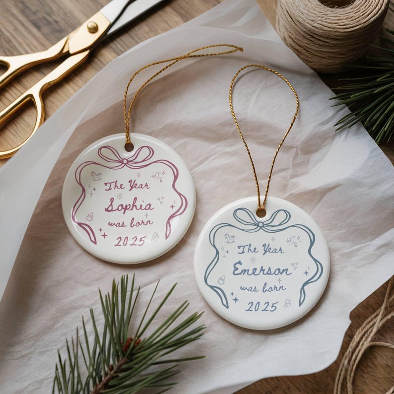 K&ouml;nnte beinhalten: Zwei wei&szlig;e runde Keramik-Weihnachtsornamente mit goldenen Kordelaufh&auml;ngern. Eines hat ein rosa Design mit dem Text "The Year Sophia was born 2025". Das andere hat ein blaues Design mit dem Text "The Year Emerson was born 2025".