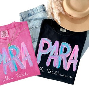 May include: Two t-shirts, one pink and one black, with the word "PARA" in a colorful, brushstroke design. The pink shirt says "Mrs. Park" and the black shirt says "Ms. Williams". A gold necklace, jeans, and a tan hat are also in the image.
