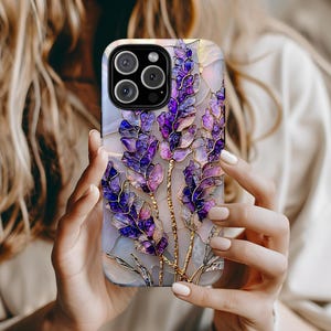 May include: A phone case featuring a lavender floral design with purple and gold accents. The case has a glossy finish and a protective design. The floral pattern is detailed and artistic, with a stained-glass effect. The case is designed for a smartphone.