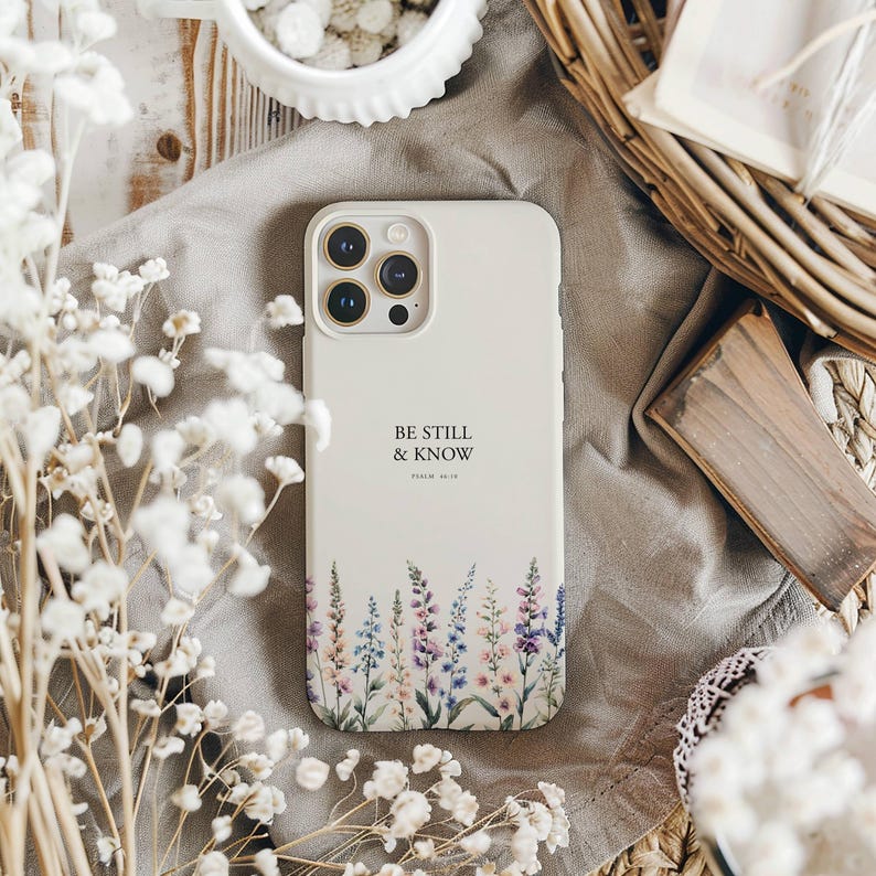 May include: A cream-colored phone case with the text "BE STILL & KNOW" and floral artwork. The case features a design of lavender, purple, and blue flowers at the bottom. The phone case is on a gray cloth with white flowers in the foreground.
