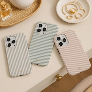 Puede incluir: Tres fundas para teléfono con patrones de rayas verticales en gris claro, verde salvia y rosa pálido. Cada funda tiene un recorte para la cámara y un monograma dorado. También se ven anillos dorados y un frasco de perfume.