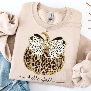 Könnte beinhalten: Beiges Sweatshirt mit einem Kürbis-Design. Der Kürbis hat einen Leopardenmuster-Körper, eine schwarz-weiß gepunktete Schleife und goldene Glitzerakzente. Der Schriftzug "hello fall" steht darunter.