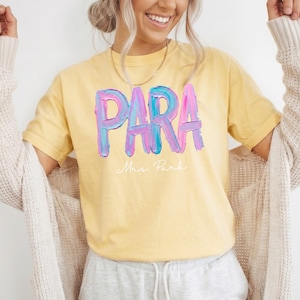 May include: Yellow t-shirt with the word "PARA" in large, colorful, brushstroke letters. Below the word is the name "Mrs. Park" in a delicate script. The shirt is a soft, casual style.