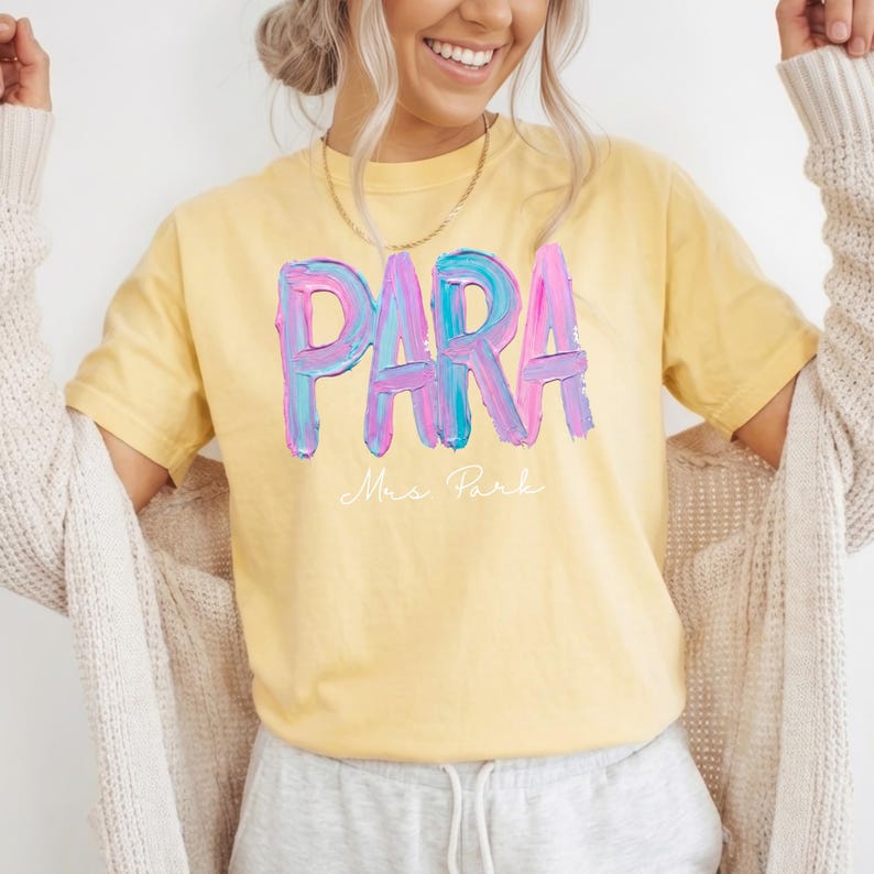 May include: Yellow t-shirt with the word "PARA" in large, colorful, brushstroke letters. Below the word is the name "Mrs. Park" in a delicate script. The shirt is a soft, casual style.