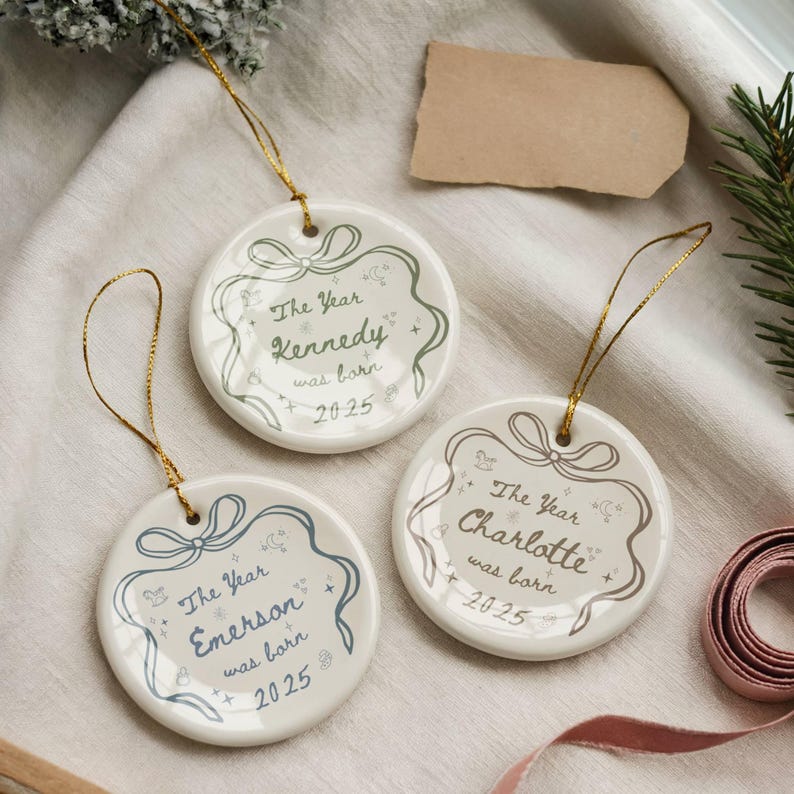 K&ouml;nnte beinhalten: Drei wei&szlig;e runde Weihnachtsornamente aus Keramik mit goldenen Aufh&auml;ngern. Jedes Ornament hat einen dekorativen Rahmen und den Text "The Year [Name] was born 2025". Die Namen sind Kennedy, Emerson und Charlotte.