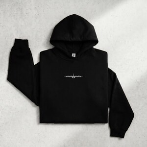May include: A black hooded sweatshirt featuring a small, white, detailed line-art graphic of a four-engine military transport aircraft centered on the chest. The garment has long sleeves, a drawstring hood, and a front kangaroo pocket.