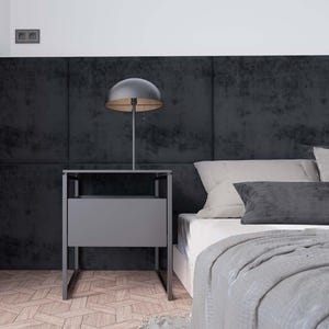 Puede incluir: Una mesita de noche negra con un caj&oacute;n y una l&aacute;mpara con una pantalla negra se encuentra junto a una cama con una cabecera gris y una manta gris.