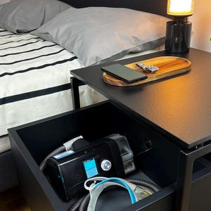 Puede incluir: Una mesita de noche negra con un caj&oacute;n abierto, que revela una m&aacute;quina CPAP y tubos. La mesa tiene una tapa negra con una bandeja de madera encima. Un tel&eacute;fono m&oacute;vil est&aacute; en la bandeja. Una l&aacute;mpara negra con una pantalla de vidrio transparente est&aacute; en la mesa.