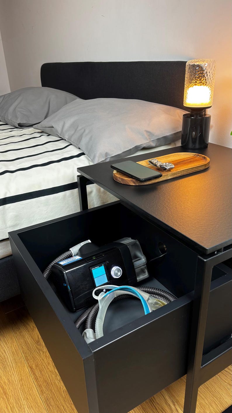 Puede incluir: Una mesita de noche negra con un caj&oacute;n abierto, que revela una m&aacute;quina CPAP y tubos. La mesa tiene una tapa negra con una bandeja de madera encima. Un tel&eacute;fono m&oacute;vil est&aacute; en la bandeja. Una l&aacute;mpara negra con una pantalla de vidrio transparente est&aacute; en la mesa.