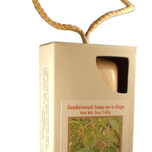 May include: A white box with a rope handle contains a bar of sandalwood soap. The box has a label that reads "Sandalwood Soap-on-a-Rope" and "Net Wt: 5oz/142g". The label also features a photo of sandalwood leaves and the text "Made by hand in the U.S.A., Mobility Bath, Waterville, Ohio".