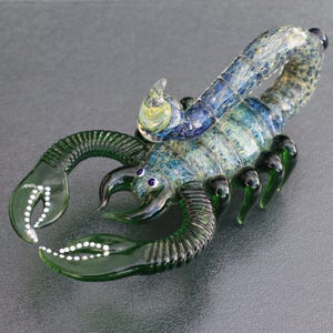 May include: A detailed glass scorpion sculpture. The scorpion features a blue and green body with black claws and legs. The claws have white dot accents. The tail curves upwards. The eyes are purple.