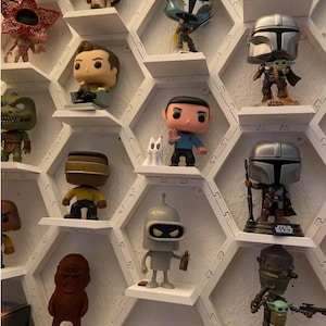 May include: A white hexagonal shelf unit with various pop culture figures displayed. The figures include characters from Star Wars, The Simpsons, and Stranger Things. The shelf unit is made of white plastic and has a honeycomb design.