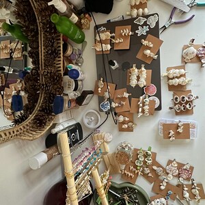 Puede incluir: Primer plano de una mesa con diversos materiales de artesanía, incluyendo conchas marinas, cuentas y pinzas para el pelo. Hay varios recipientes de cuentas y una variedad de pinzas para el pelo con conchas marinas adheridas. La mesa está cubierta con un mantel blanco.