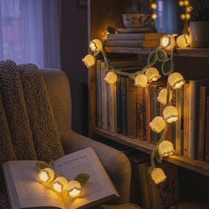 May include: A warm, inviting scene with a crochet flower garland illuminated against a bookshelf and an open book. The garland's soft light highlights the room, with a blanket and armchair in the foreground. The book's title is "The Herbarium's Song".