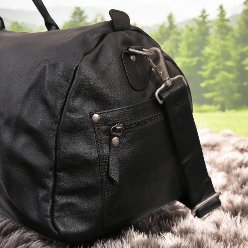Puede incluir: Bolsa de viaje de cuero negro con un bolsillo con cremallera y una correa para el hombro. La bolsa tiene un cierre de metal plateado.