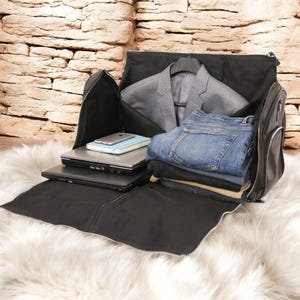 Könnte beinhalten: Eine schwarze Leder-Kleidertasche mit Reißverschlussöffnung, die einen Anzug, Jeans und einen Laptop im Inneren zeigt.