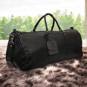 Puede incluir: Una bolsa de viaje de cuero negro con asa superior y una etiqueta de equipaje desmontable. La bolsa est&aacute; sobre una alfombra peluda de color marr&oacute;n.