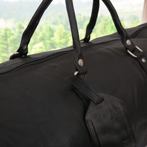 Puede incluir: Bolsa de viaje de cuero negro con herrajes plateados y cierre de cremallera. La bolsa tiene dos asas superiores y una etiqueta de cuero desmontable.
