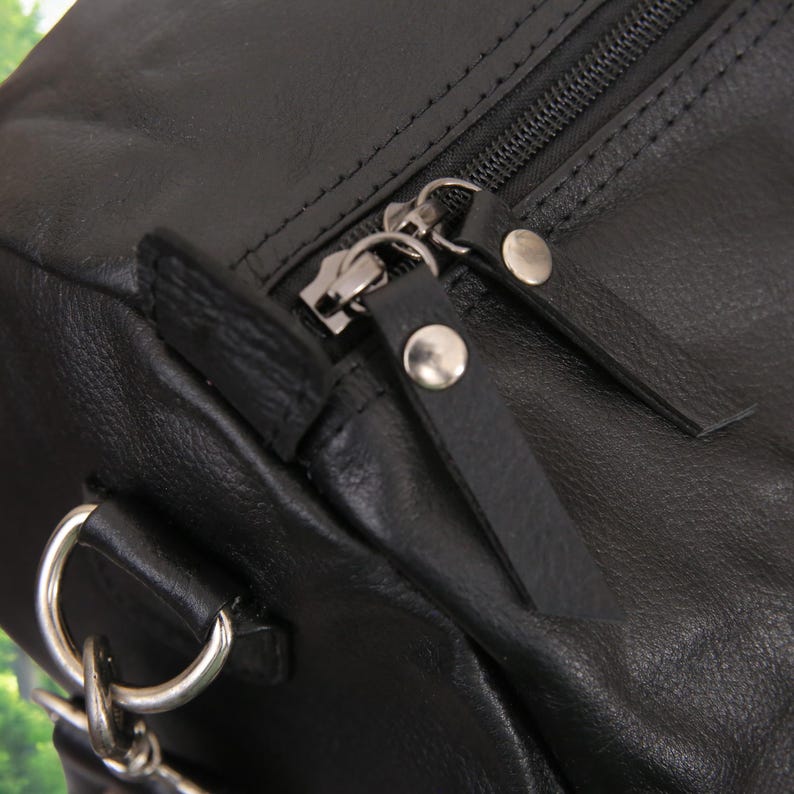 Puede incluir: Primer plano de una bolsa de viaje de cuero negro con cierre de cremallera y herrajes plateados. La bolsa tiene una correa con una hebilla de metal.