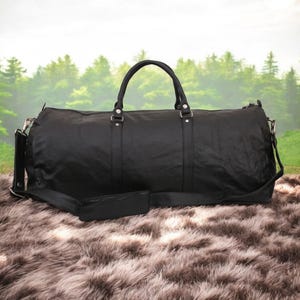 Puede incluir: Una bolsa de viaje de cuero negro con una correa de hombro larga y dos asas superiores. La bolsa est&aacute; sobre una superficie de piel sint&eacute;tica marr&oacute;n.