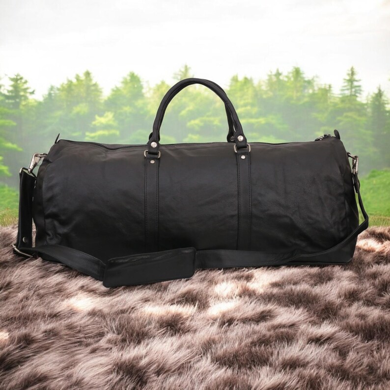Puede incluir: Una bolsa de viaje de cuero negro con una correa de hombro larga y dos asas superiores. La bolsa est&aacute; sobre una superficie de piel sint&eacute;tica marr&oacute;n.
