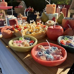 Op de afbeelding: Een display van handgemaakte kaarsen in de vorm van voedsel. De kaarsen omvatten een wafel, appels, peren en kommen met ontbijtgranen. De kaarsen zijn felgekleurd en gerangschikt op een houten dienblad, wat een unieke uitstraling creëert.