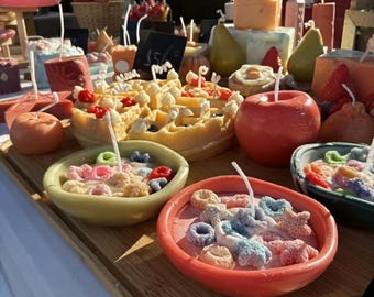 May include: A display of handmade candles shaped like food items. The candles include a waffle, apples, pears, and bowls of cereal. The candles are brightly colored and arranged on a wooden tray, creating a whimsical and unique aesthetic.