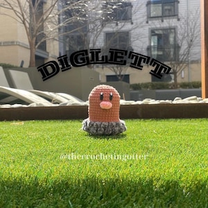 May include: A crocheted Diglett Pokemon plush toy with a brown and pink body and a gray head. The toy is sitting on a green grassy surface with the text "DIGLETT" in black letters above it.