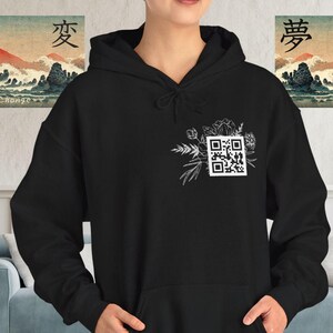 Puede incluir: Sudadera con capucha negra con un diseño floral blanco y un código QR en el pecho. La sudadera tiene una capucha con cordón y un bolsillo de canguro. Dos pinturas de paisajes enmarcadas con caracteres japoneses están en el fondo.