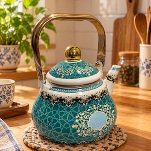 May include: A teal enamel tea kettle featuring a geometric pattern, a gold-toned metal handle, and a matching lid with a gold-toned knob. The kettle has a white spout and a decorative border with intricate floral details in black, white, and gold.