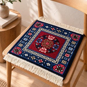 May include: A square rug with a deep red centre featuring a circular floral motif and a geometric border in blue, black and white. The rug has a white fringe on two opposite sides and is placed on a wooden chair seat, displaying intricate patterns.