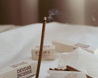 May include: A burning Devdar incense stick with wisps of smoke, placed in a black circular holder. Several incense sticks and boxes of Devdar Incense are visible. The packaging is white with black text.