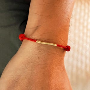 May include: A red and gold bracelet is worn on a wrist. The bracelet is made of red string with a gold-colored central section. The bracelet is tied with knots at each end.
