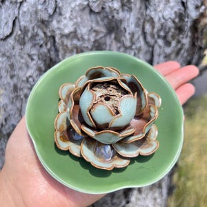 May include: A ceramic incense holder shaped like a lotus flower. The holder features a light green dish with a detailed flower in brown and blue hues. The incense holder is held in a hand.