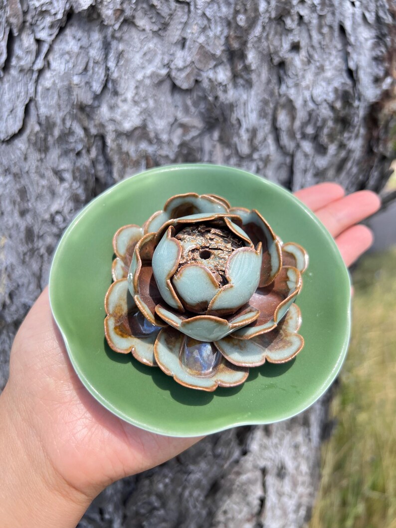 May include: A ceramic incense holder shaped like a lotus flower. The holder features a light green dish with a detailed flower in brown and blue hues. The incense holder is held in a hand.