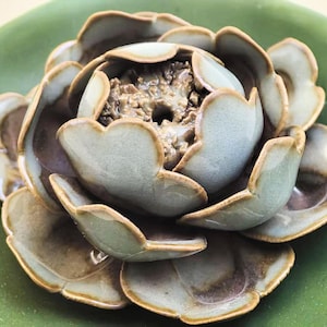 May include: A ceramic lotus flower sculpture in shades of green and brown, resting on a green plate. The petals have a textured finish and are arranged in layers, creating a detailed floral design.