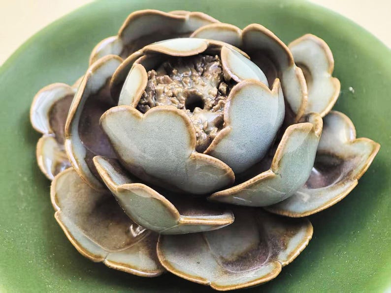 May include: A ceramic lotus flower sculpture in shades of green and brown, resting on a green plate. The petals have a textured finish and are arranged in layers, creating a detailed floral design.