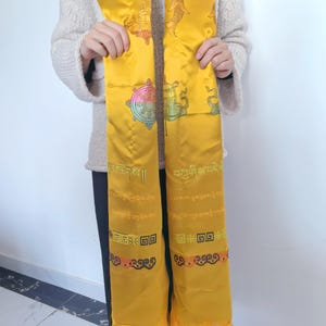 Könnte beinhalten: Ein gelber Seidenschal mit aufwendigen Stickereien von buddhistischen Symbolen und Mantras. Der Schal hat einen Fransenbesatz am unteren Rand und ist mit bunten Mustern verziert.