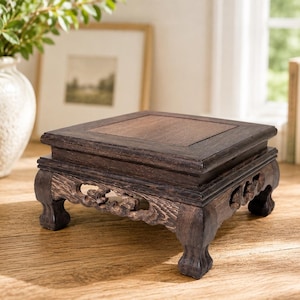 May include: A square dark brown wooden display stand featuring a flat top surface and four carved legs with intricate openwork detailing. The stand has a visible wood grain texture and a raised border around the central square panel.