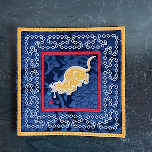 May include: A square fabric patch featuring a central embroidered golden tiger on a dark blue floral background. The design is framed by a red border, a white geometric chain pattern, and an outer yellow stitched edge on a dark blue fabric.
