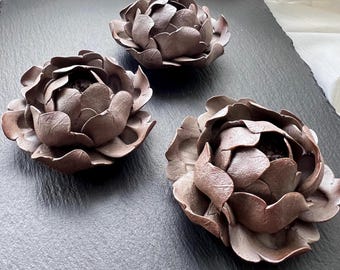 May include: Three brown, flower-shaped decorative bowls with textured petals are arranged on a dark grey surface. The bowls have a detailed, organic design, resembling peonies. The lighting highlights the texture and form of the bowls.