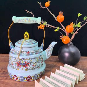 May include: A light blue ceramic teapot with a floral design and gold accents. The teapot has a curved spout, a handle, and a lid with a gold finial. A small gold chain connects the lid to the handle. A vase with orange fruit and a book are also in the image.