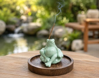 May include: A light green ceramic frog incense holder sits on a circular dark brown wooden tray. A thin incense stick is inserted into the frog, with a visible trail of white smoke rising upward from the lit tip of the incense stick.