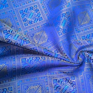 Könnte beinhalten: Nahaufnahme eines leuchtend blauen Stoffes mit einem geometrischen Muster. Das Design zeigt Quadrate und Rechtecke mit komplizierten Details in Blau- und Goldtönen. Der Stoff hat einen leichten Glanz, was auf ein luxuriöses Material hindeutet.