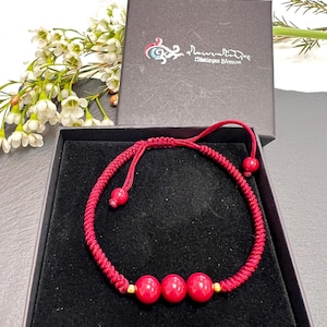 May include: A red string bracelet with three red beads and gold accents. The bracelet is in a black box with a white logo that says "Rituals & Woven".